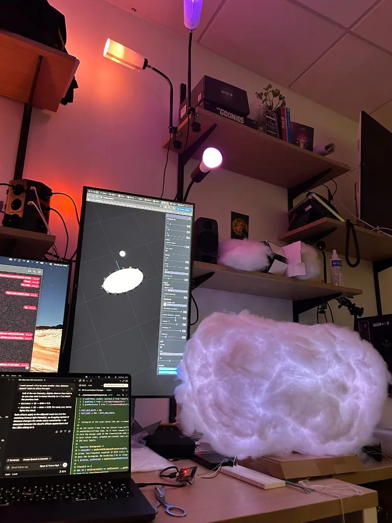 A small illuminated cloud prototype beside computers running a three-dimensional simulation